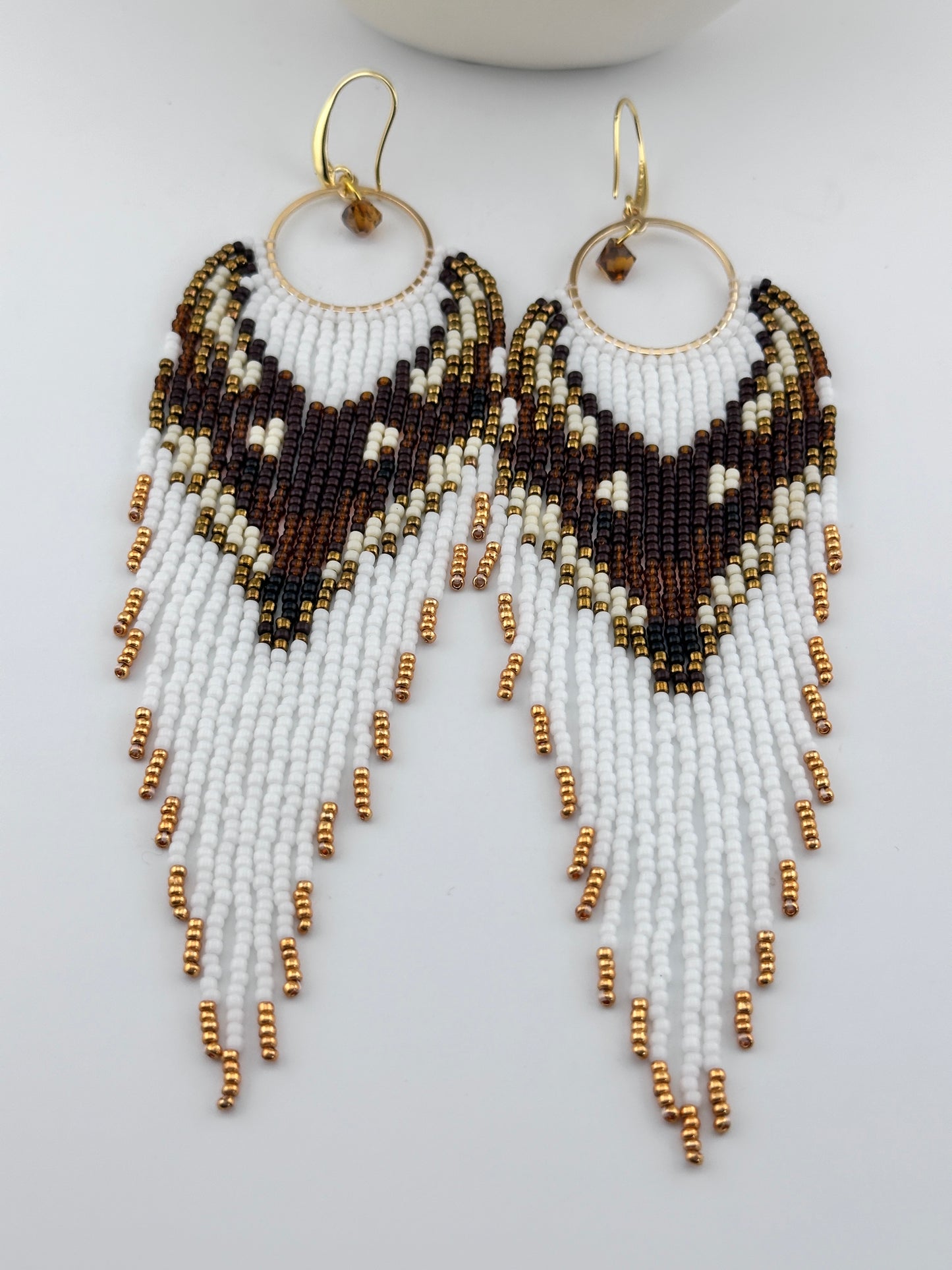 Handmade beaded hoop earrings with gold hooks, featuring white, brown, black, and gold seed beads in a chevron fringe pattern, displayed on a white background.