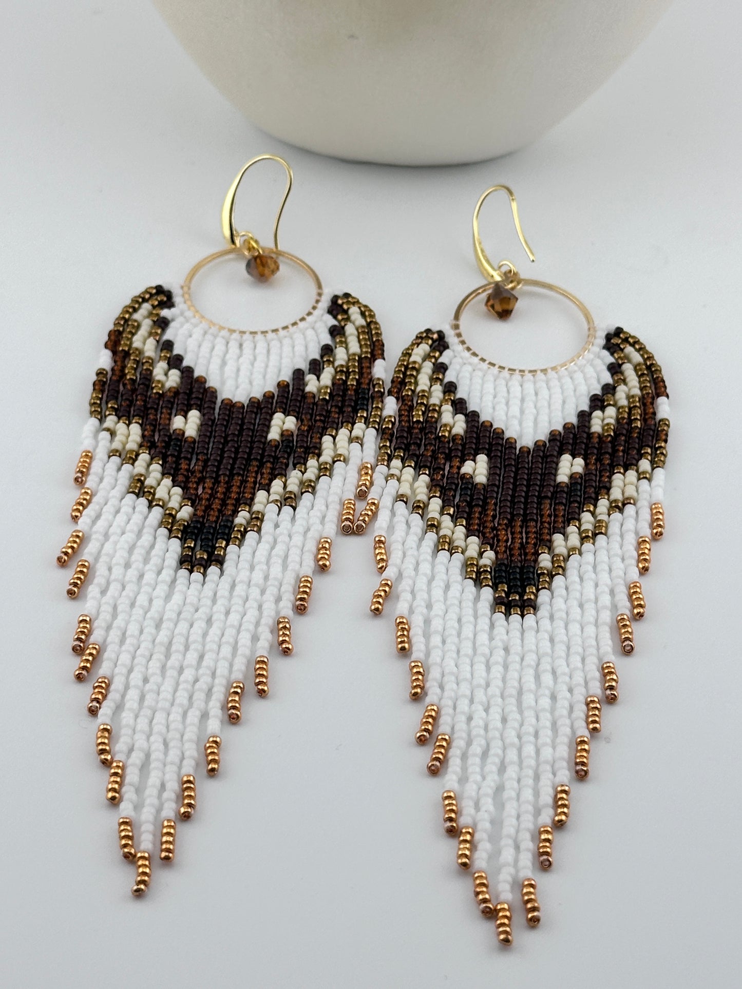 Handmade beaded hoop earrings with gold hooks, featuring white, brown, black, and gold seed beads in a chevron fringe pattern, displayed on a white background.