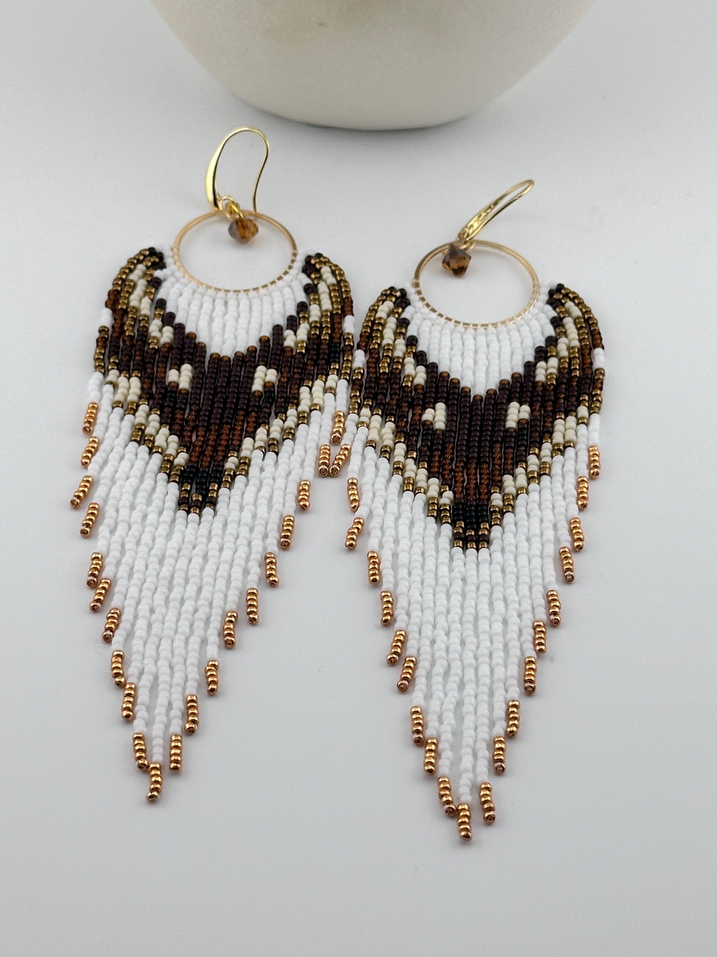 Handmade beaded hoop earrings with gold hooks, featuring white, brown, black, and gold seed beads in a chevron fringe pattern, displayed on a white background.