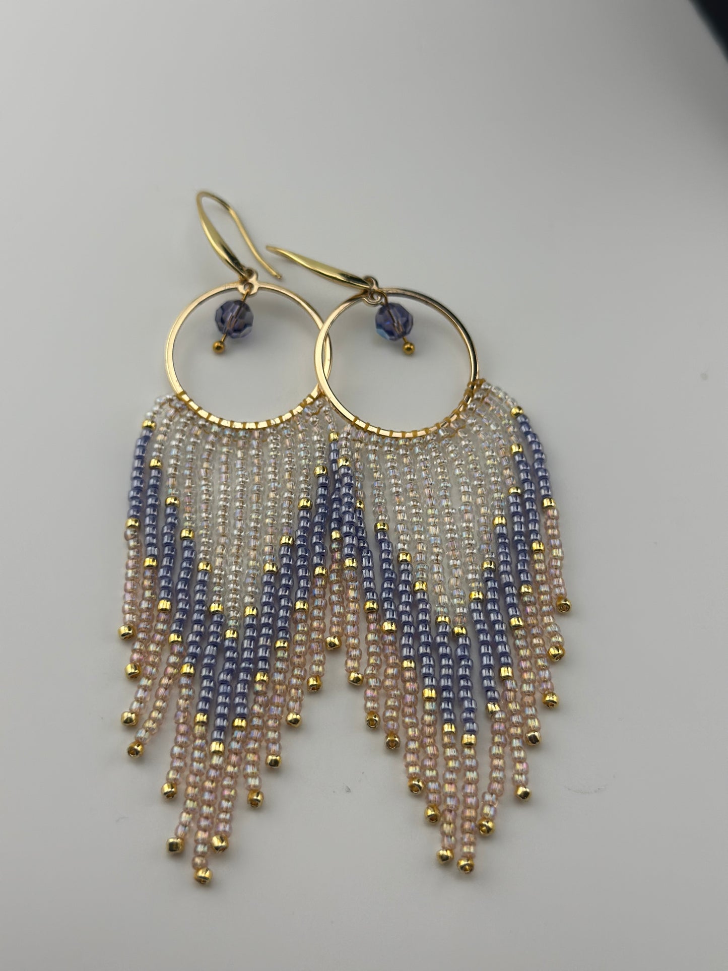Handmade gold hoop earrings with soft pink, lavender, and pearl white seed bead fringe in a chevron pattern, finished with gold accents and purple crystal dangles