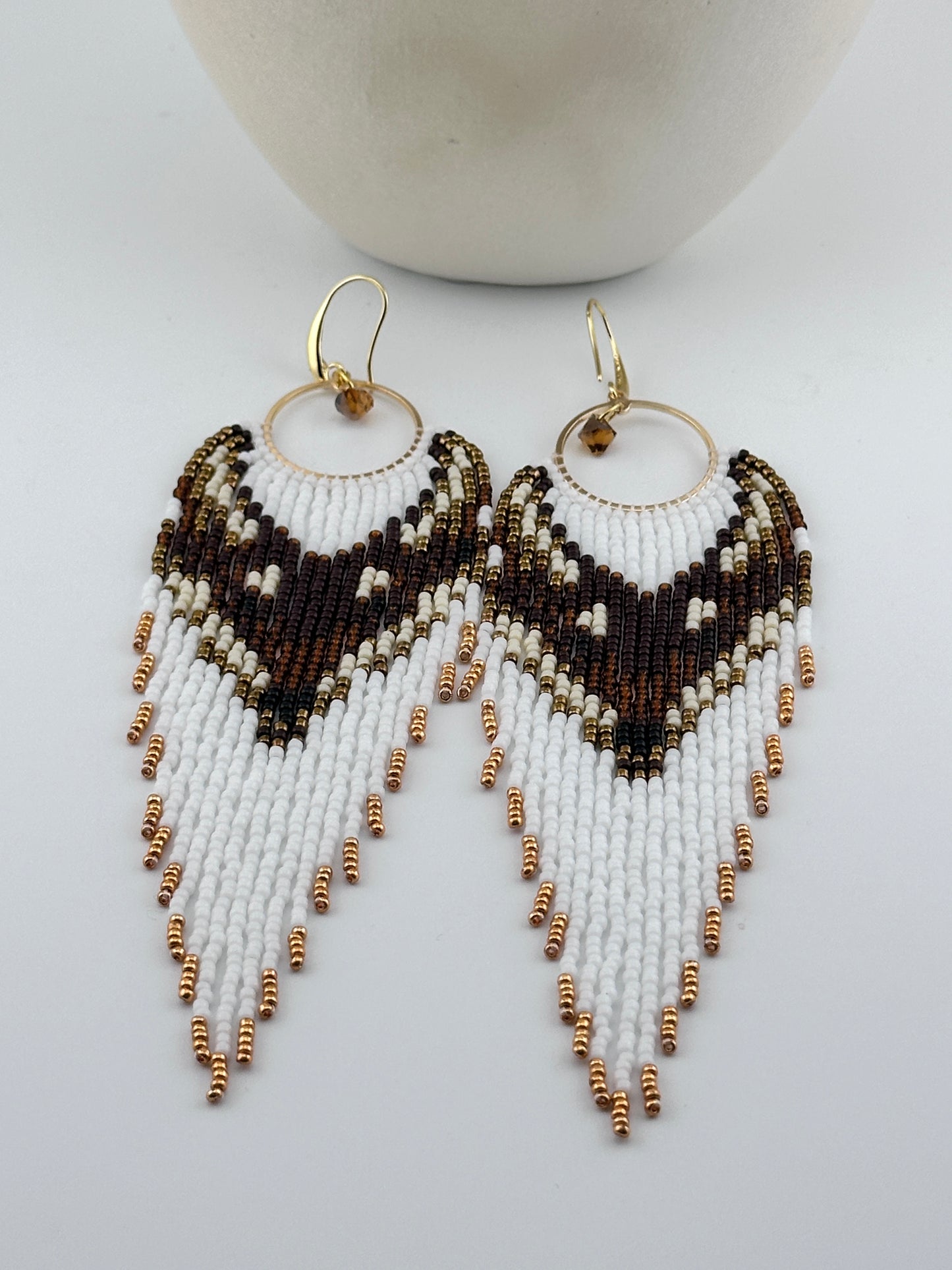 Handmade beaded hoop earrings with gold hooks, featuring white, brown, black, and gold seed beads in a chevron fringe pattern, displayed on a white background.