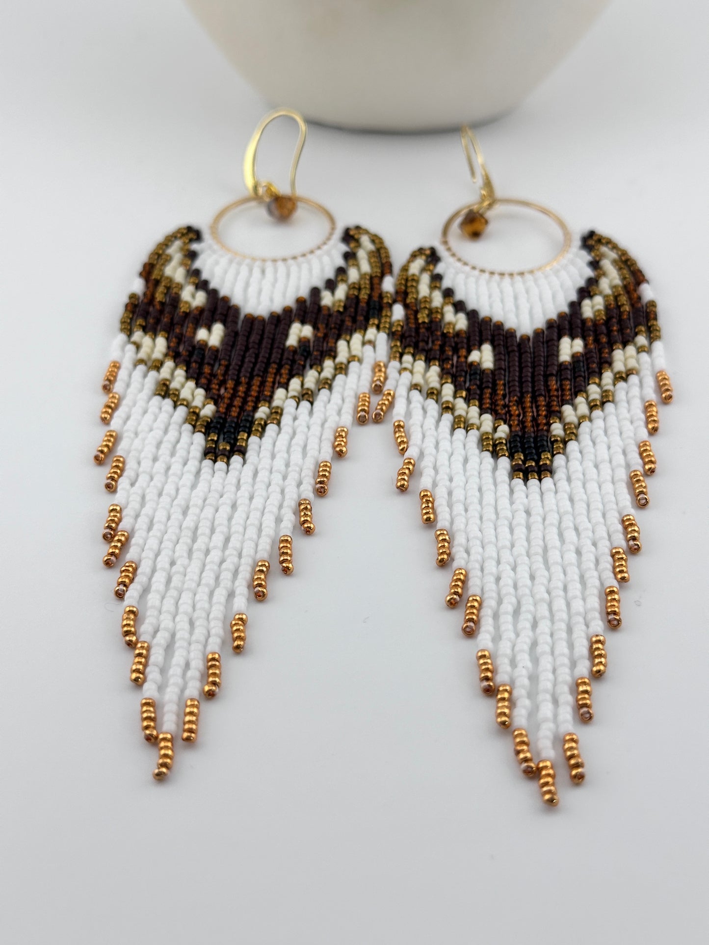 Handmade beaded hoop earrings with gold hooks, featuring white, brown, black, and gold seed beads in a chevron fringe pattern, displayed on a white background.