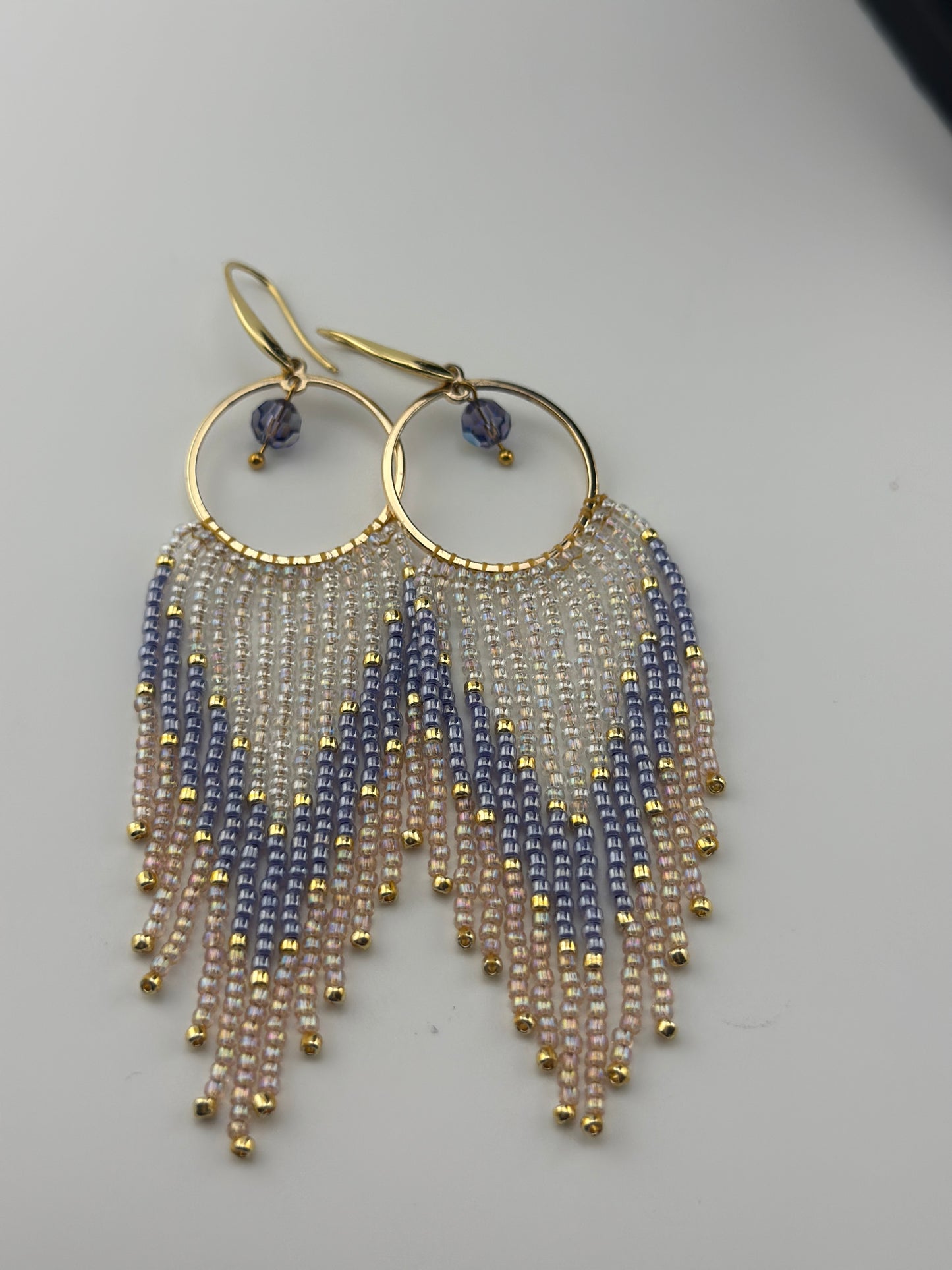 Handmade gold hoop earrings with soft pink, lavender, and pearl white seed bead fringe in a chevron pattern, finished with gold accents and purple crystal dangles