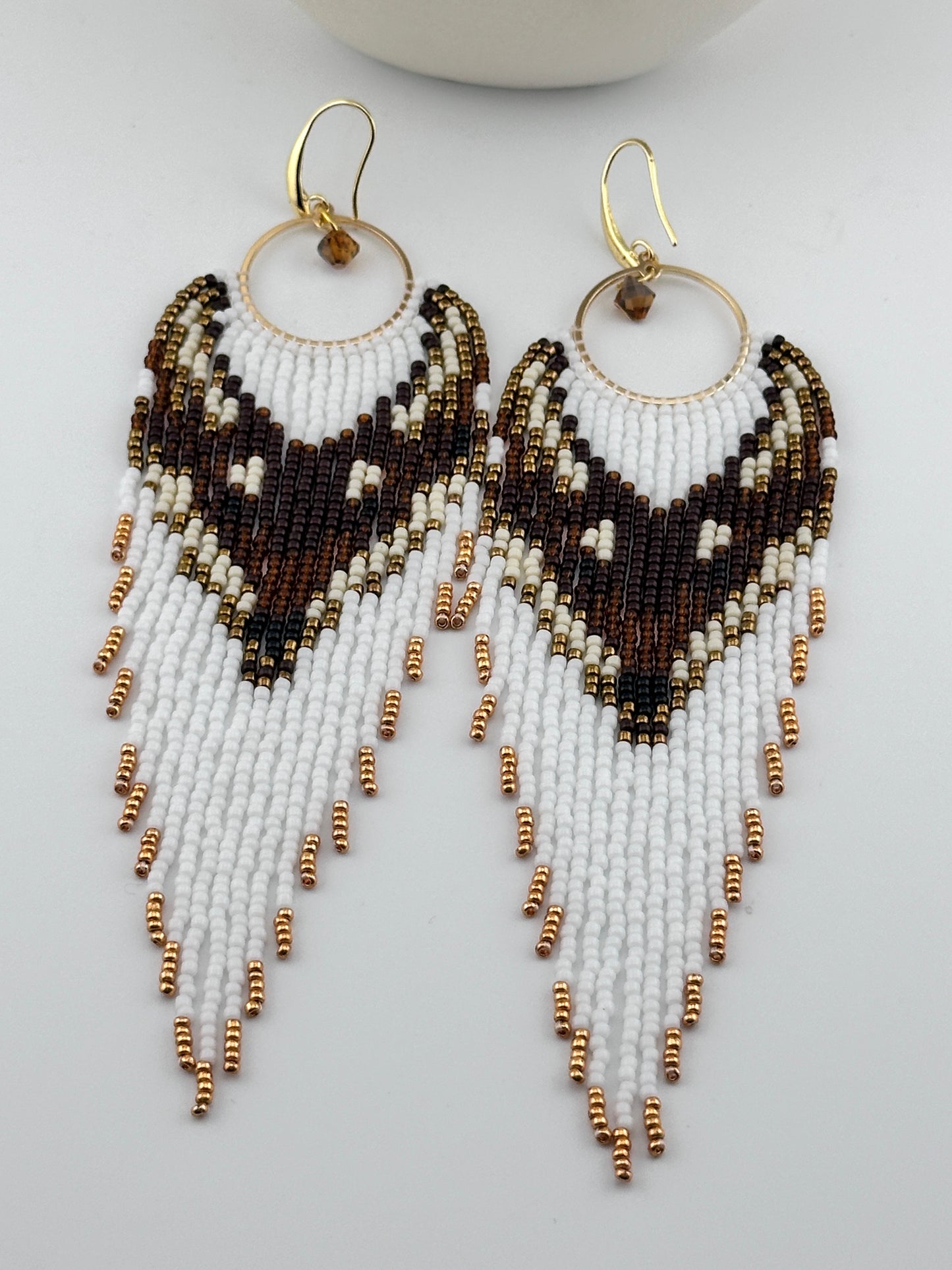 
Handmade beaded hoop earrings with gold hooks, featuring white, brown, black, and gold seed beads in a chevron fringe pattern, displayed on a white background.