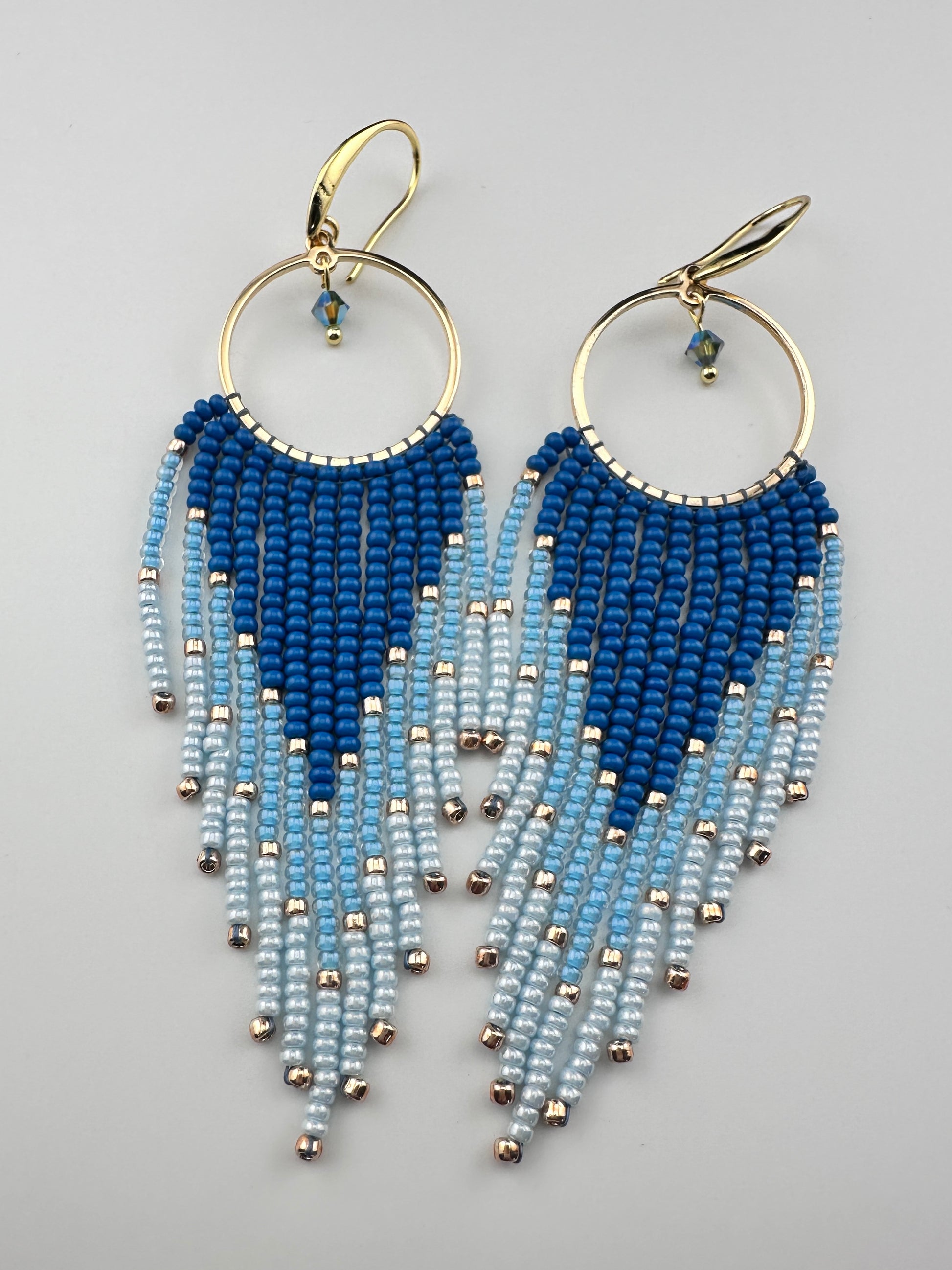 
Handmade gold hoop earrings with ombré blue Miyuki seed beads in a long fringe design, laid flat on a soft white background.
