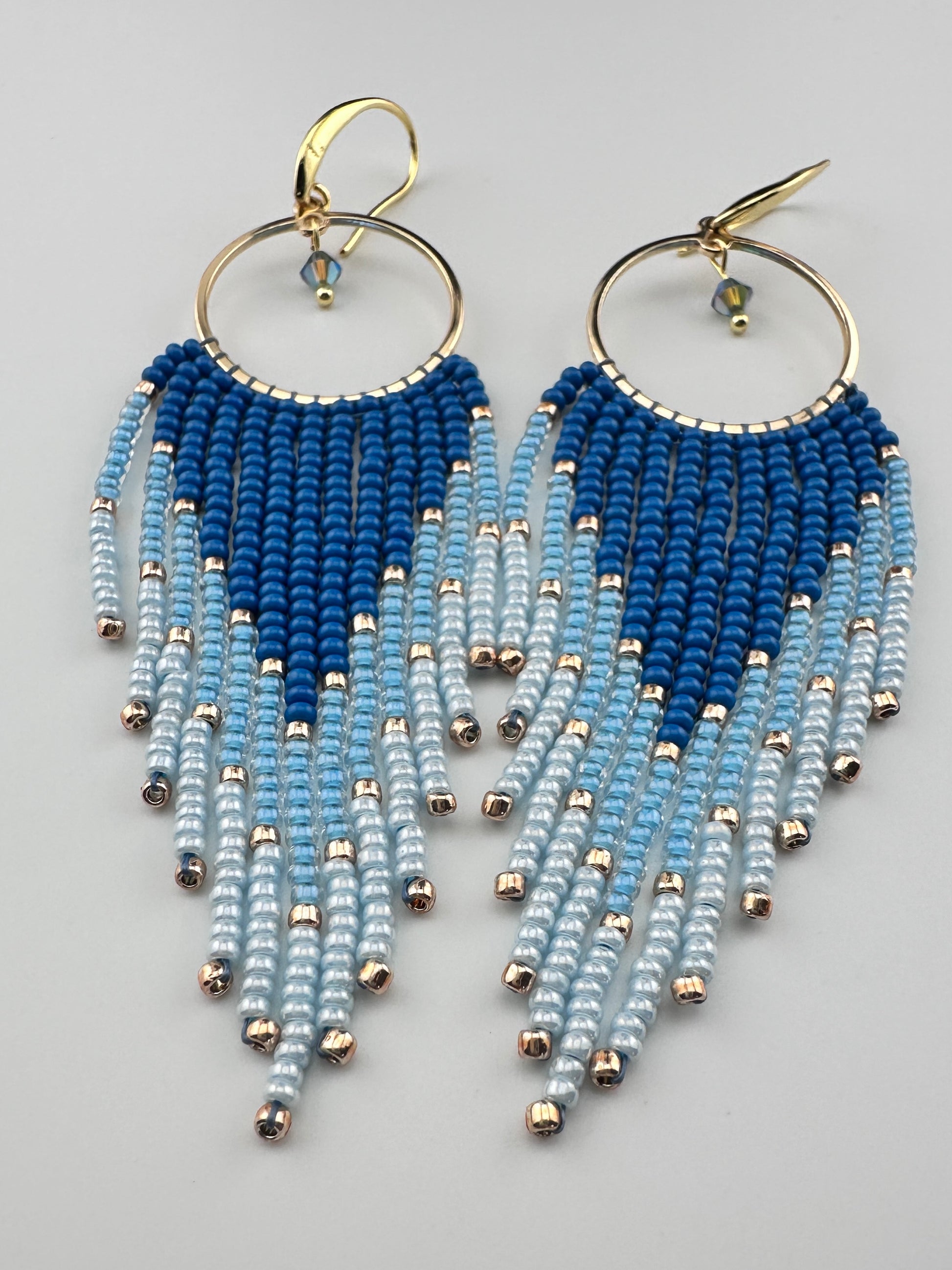 
Handmade gold hoop earrings with ombré blue Miyuki seed beads in a long fringe design, laid flat on a soft white background.
