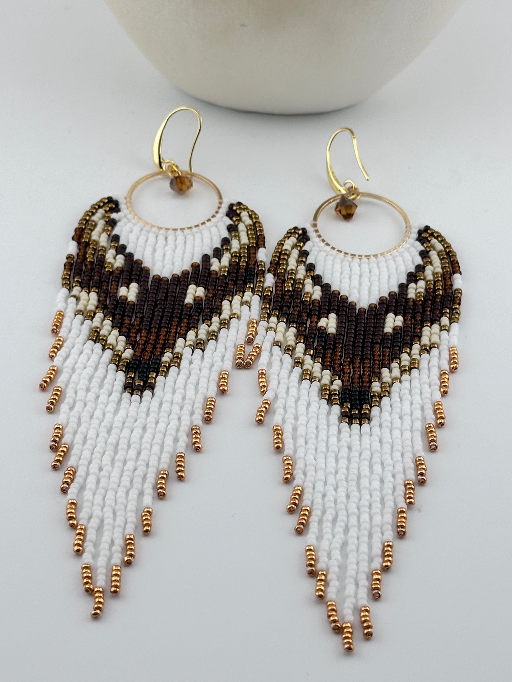 
Handmade beaded hoop earrings with gold hooks, featuring white, brown, black, and gold seed beads in a chevron fringe pattern, displayed on a white background.