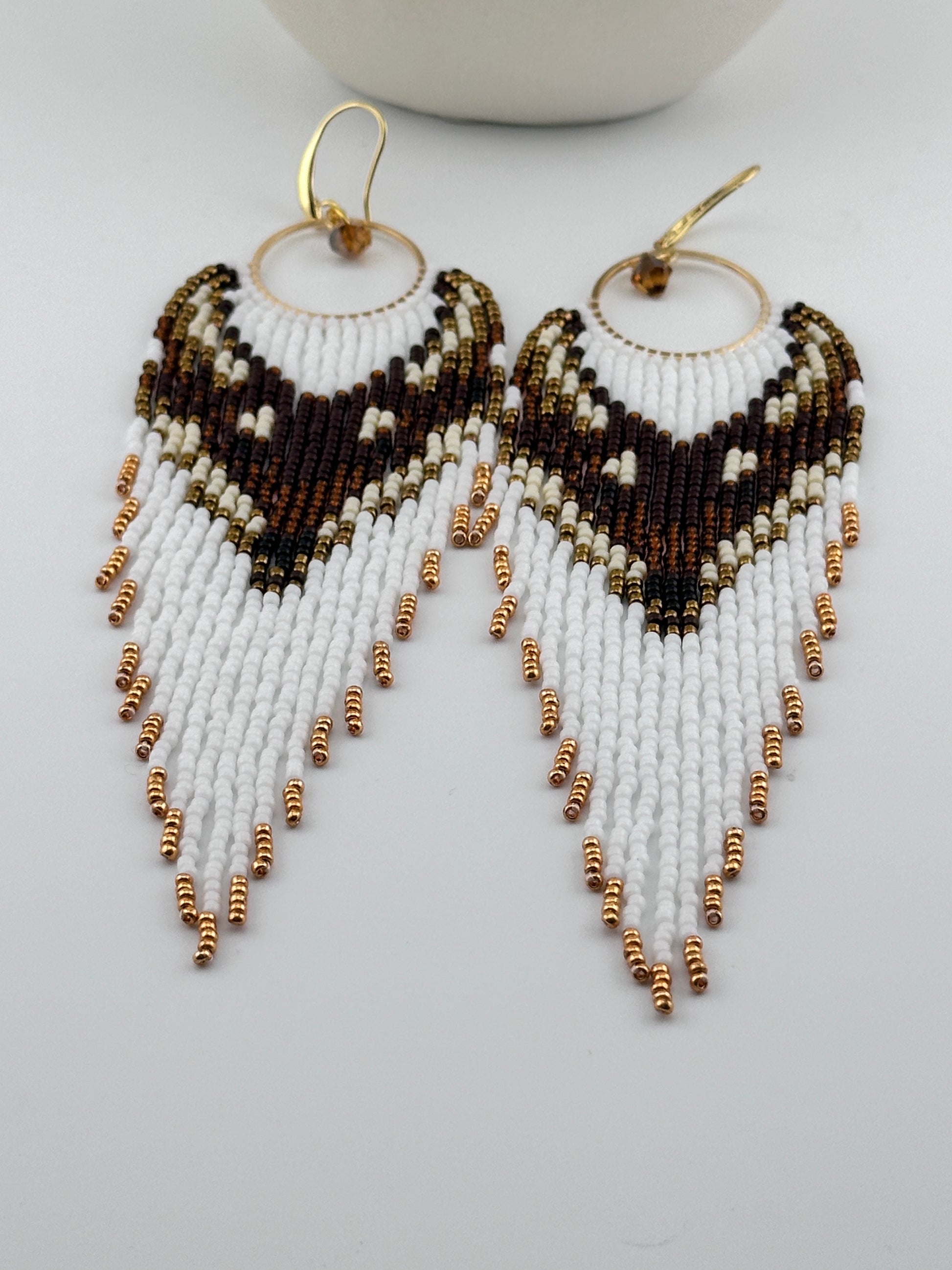 
Handmade beaded hoop earrings with gold hooks, featuring white, brown, black, and gold seed beads in a chevron fringe pattern, displayed on a white background.