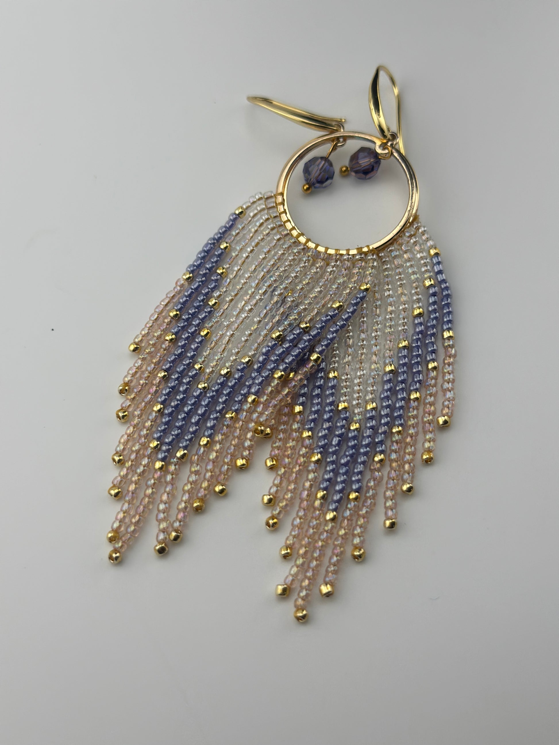 Handmade gold hoop earrings with soft pink, lavender, and pearl white seed bead fringe in a chevron pattern, finished with gold accents and purple crystal dangles
