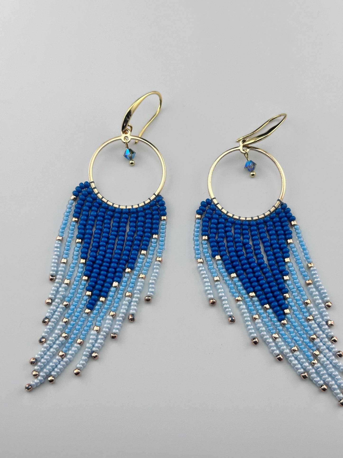 
Handmade gold hoop earrings with ombré blue Miyuki seed beads in a long fringe design, laid flat on a soft white background.
