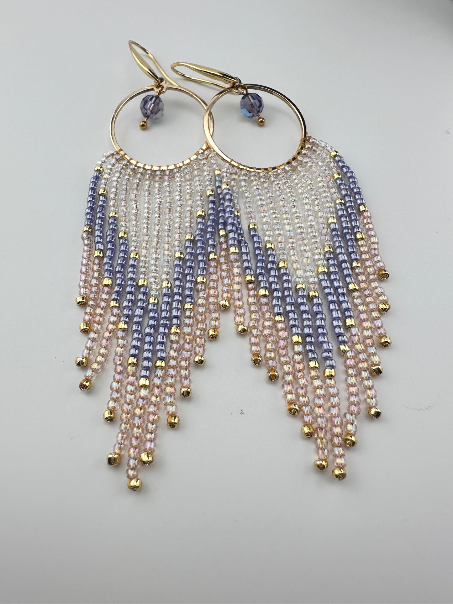 Handmade gold hoop earrings with soft pink, lavender, and pearl white seed bead fringe in a chevron pattern, finished with gold accents and purple crystal dangles