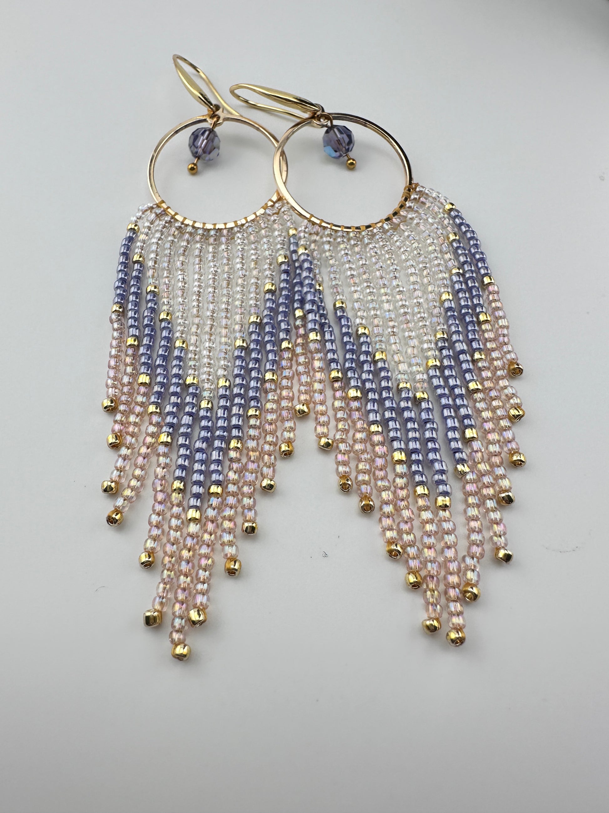 Handmade gold hoop earrings with soft pink, lavender, and pearl white seed bead fringe in a chevron pattern, finished with gold accents and purple crystal dangles