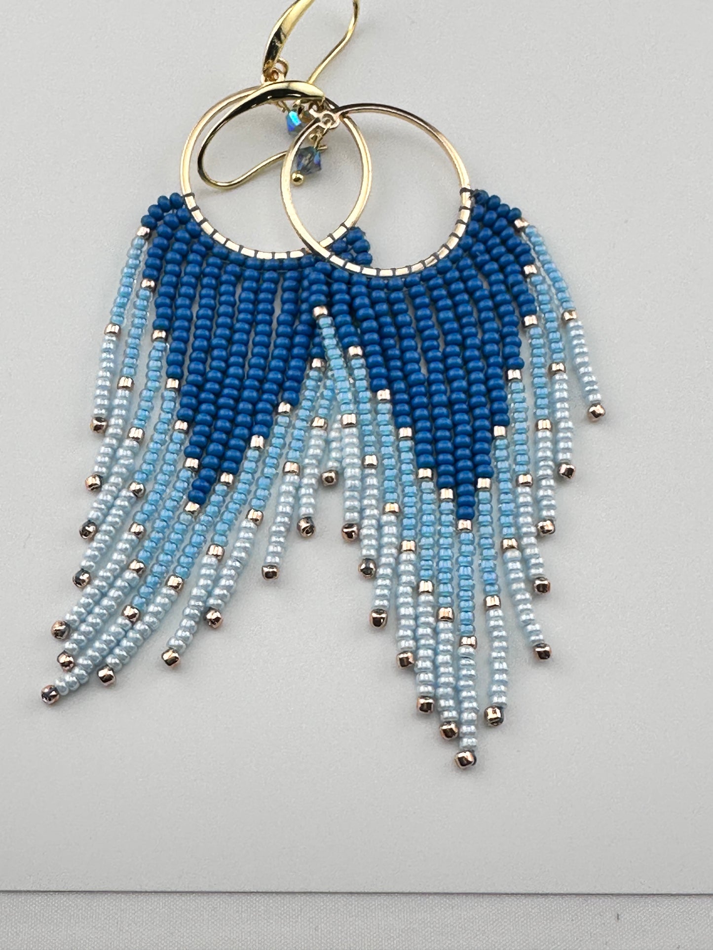
Handmade gold hoop earrings with ombré blue Miyuki seed beads in a long fringe design, laid flat on a soft white background.
