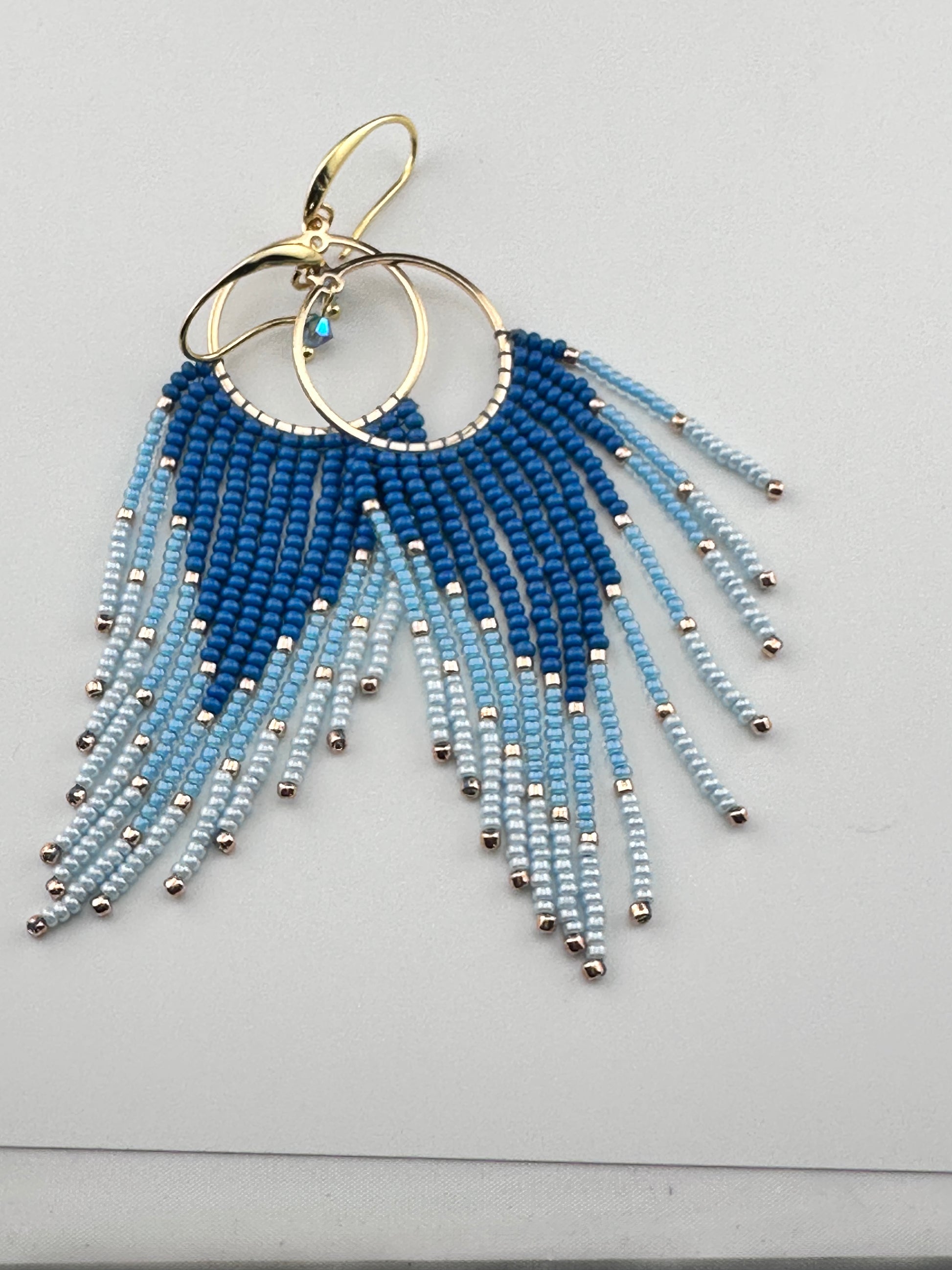 
Handmade gold hoop earrings with ombré blue Miyuki seed beads in a long fringe design, laid flat on a soft white background.
