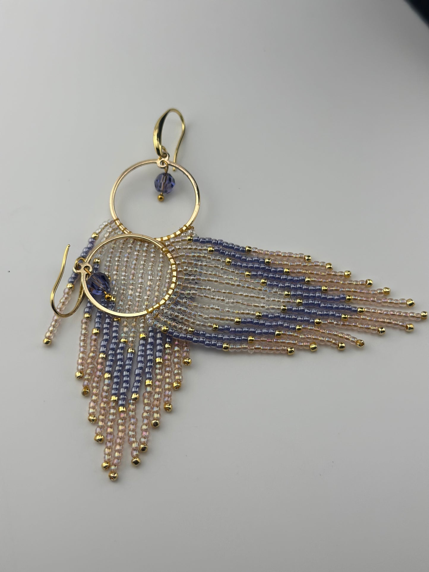 Handmade gold hoop earrings with soft pink, lavender, and pearl white seed bead fringe in a chevron pattern, finished with gold accents and purple crystal dangles