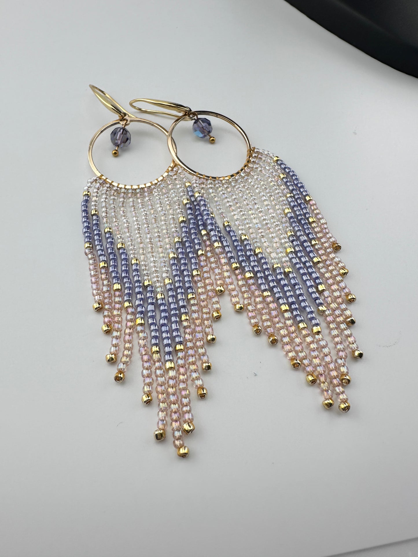 Handmade gold hoop earrings with soft pink, lavender, and pearl white seed bead fringe in a chevron pattern, finished with gold accents and purple crystal dangles