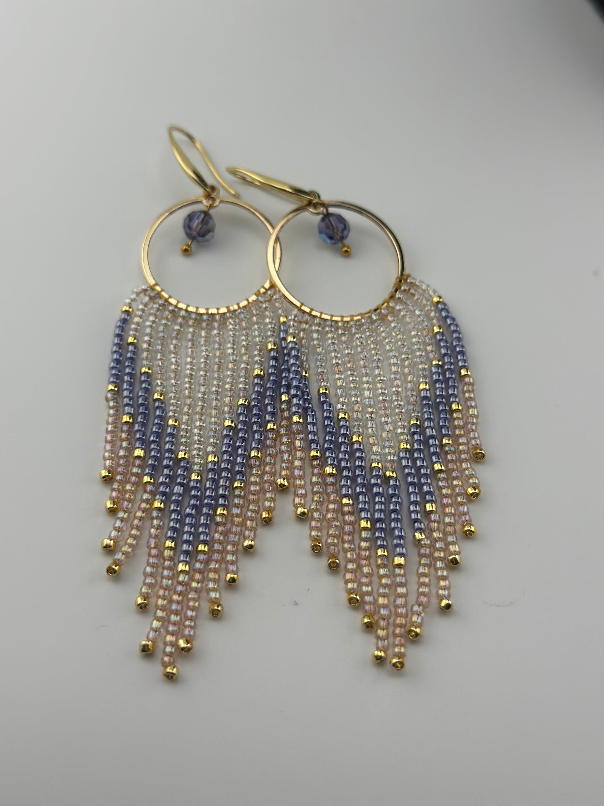 Handmade gold hoop earrings with soft pink, lavender, and pearl white seed bead fringe in a chevron pattern, finished with gold accents and purple crystal dangles