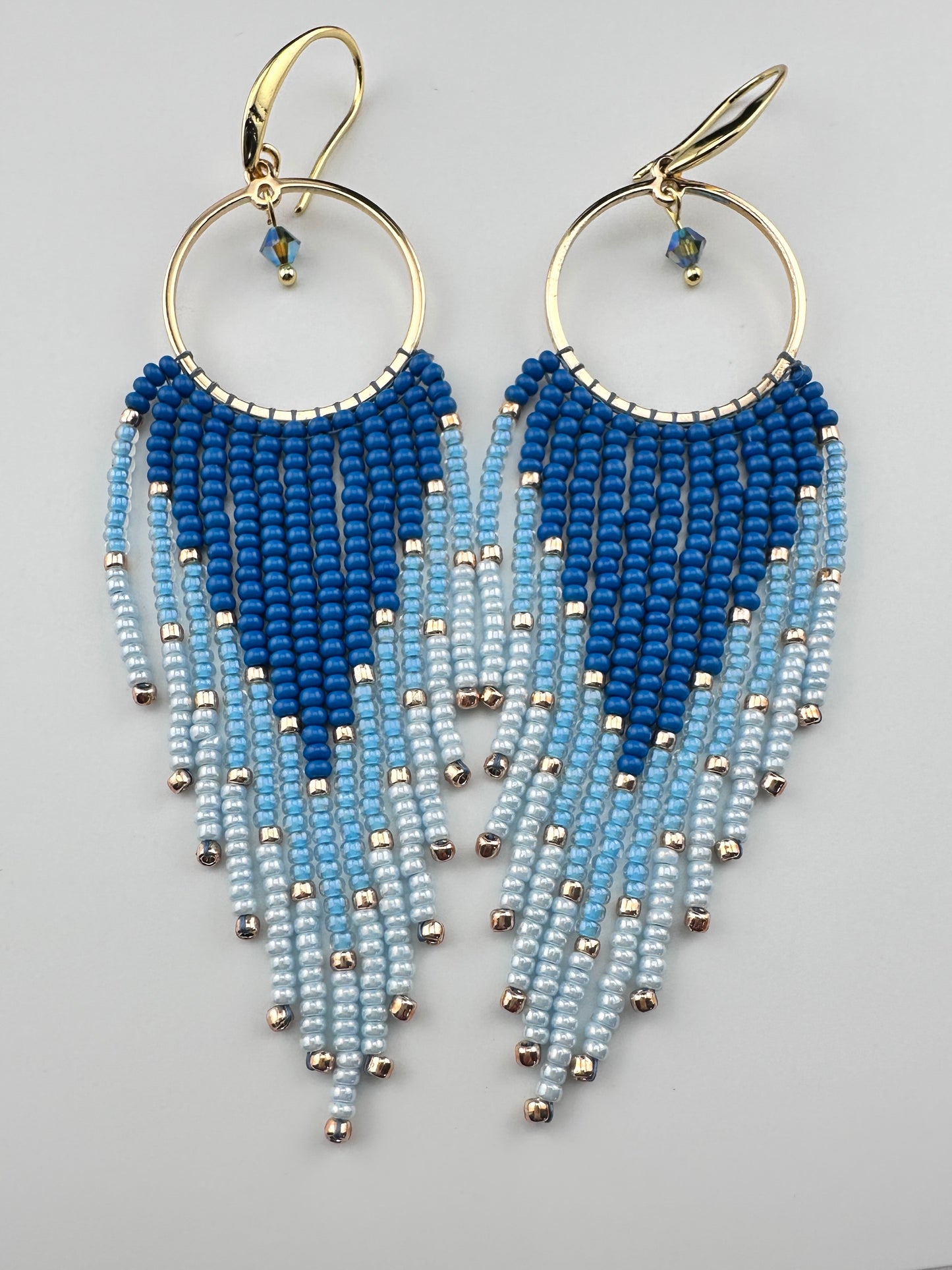 
Handmade gold hoop earrings with ombré blue Miyuki seed beads in a long fringe design, laid flat on a soft white background.
