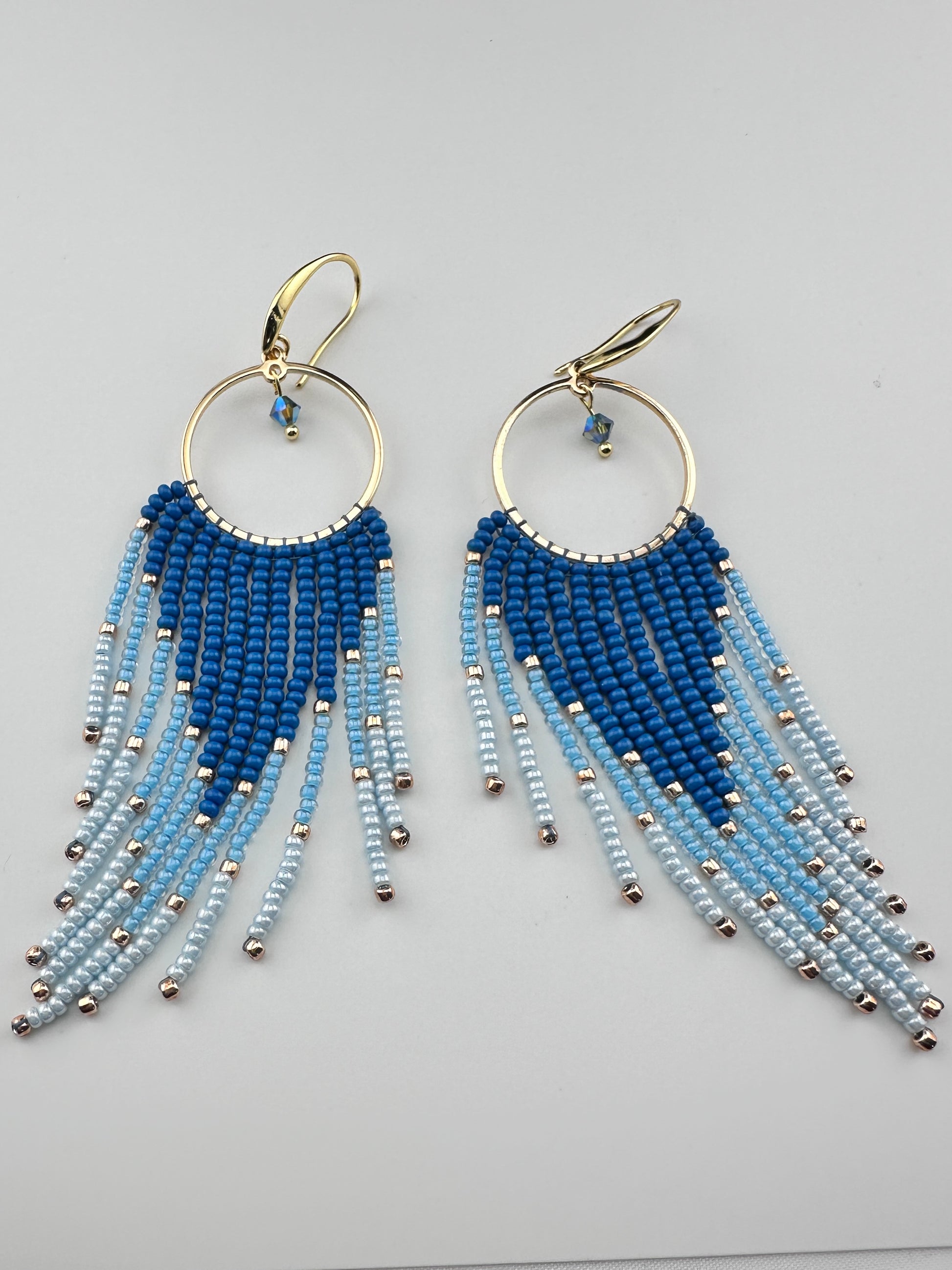 
Handmade gold hoop earrings with ombré blue Miyuki seed beads in a long fringe design, laid flat on a soft white background.
