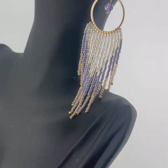 Handmade gold hoop earrings with soft pink, lavender, and pearl white seed bead fringe in a chevron pattern, finished with gold accents and purple crystal dangles