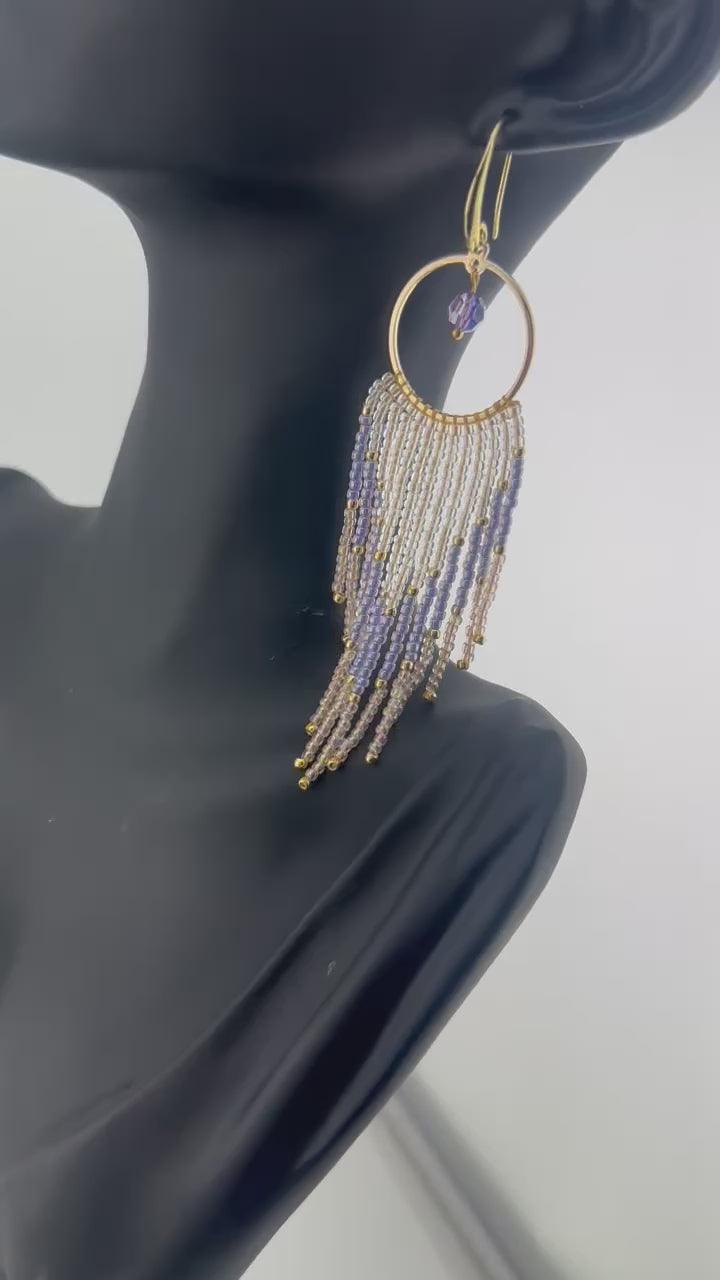 Handmade gold hoop earrings with soft pink, lavender, and pearl white seed bead fringe in a chevron pattern, finished with gold accents and purple crystal dangles